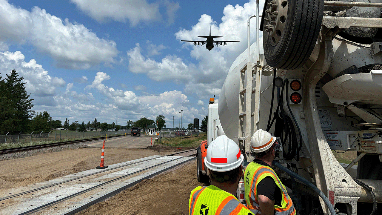 Construction workers near a concrete mixer watch a large aircraft flying overhead at a work site beside railroad tracks.