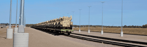 A long line of military vehicles loaded on rail cars at a rail yard under a clear sky.