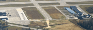 An aerial view of an airport showing intersecting runways, taxiways, hangars, and nearby buildings.