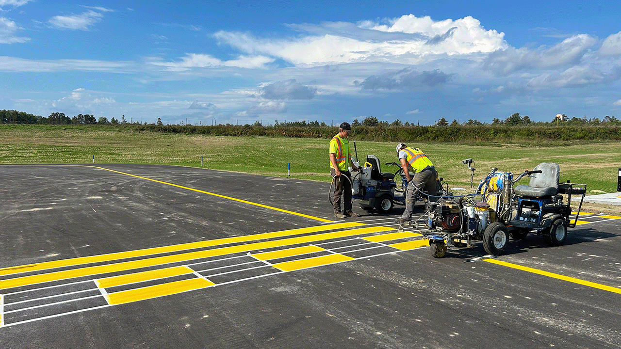 Workers painting yellow and white markings on a paved runway at Longville Airport.