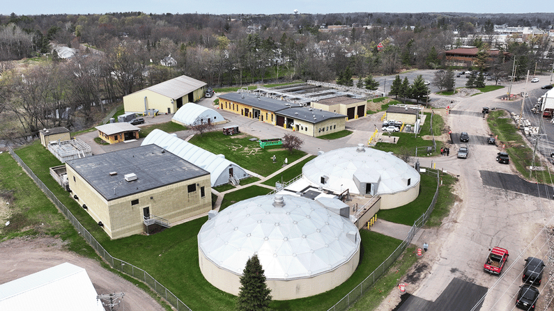 Aerial view of Medford's wastewater treatment plant with tanks, buildings, and surrounding roads, set in a green landscape near residential areas.