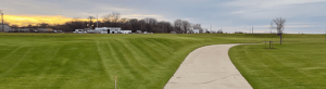 Banner image of a curving concrete path through a grassy field under an overcast sky, with trees and parked cars in the distance.