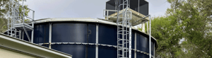Large blue cylindrical water storage tank with exterior ladder and platform beside a utility building, surrounded by trees.